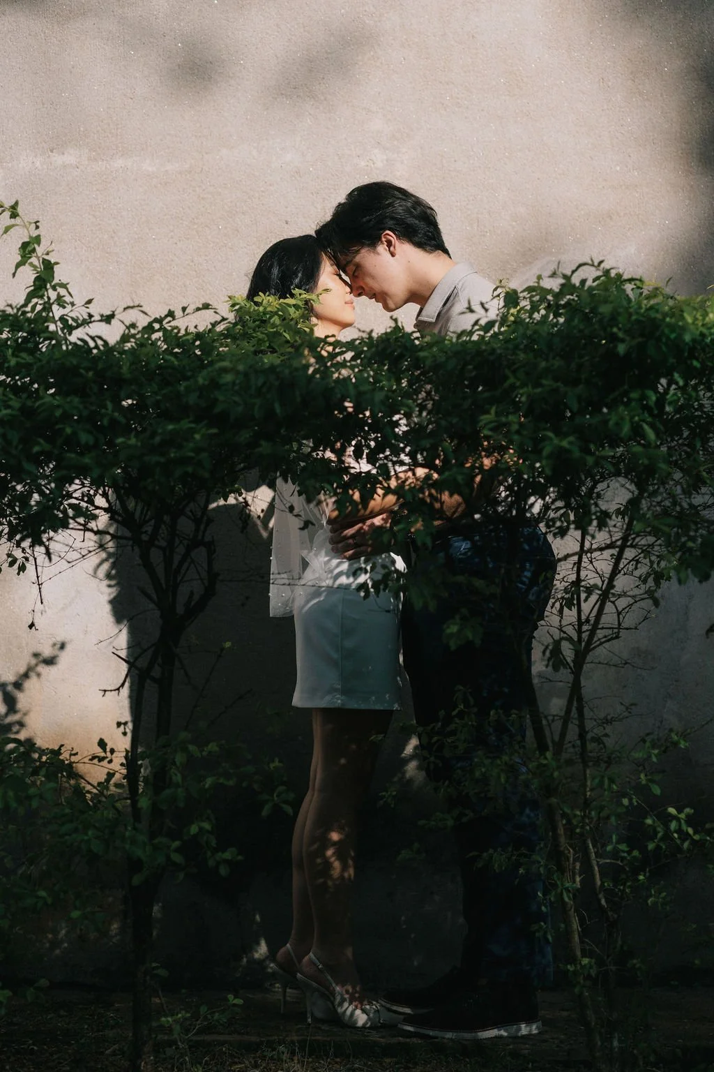 Casal se beijando com cabeça tocando, entre arbustos verdes, em ambiente ao ar livre