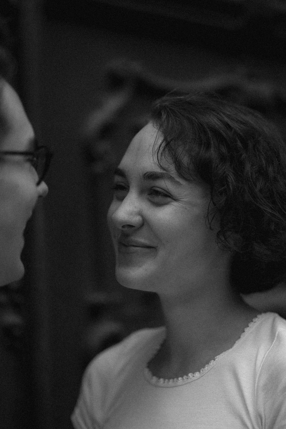 Duas pessoas conversando de perto e sorrindo, uma delas usando óculos e a outra com cabelos cacheados, em ambiente escuro.