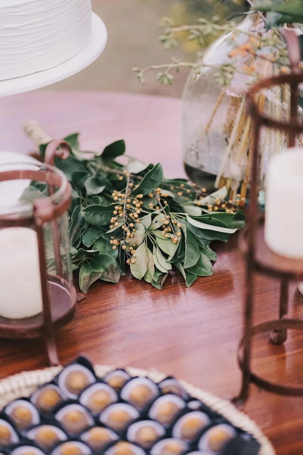 Arranjo de folhas verdes e pequenos galhos com frutos amarelos em uma mesa de madeira, com velas em vidro ao redor.