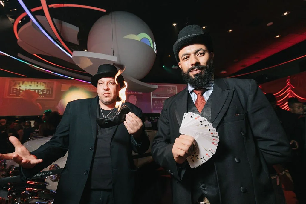 Dois homens com roupas formais e chapéus, um segurando um revólver com fogo e o outro exibindo um maço de cartas, em um ambiente com iluminação de palco e decoração futurista.