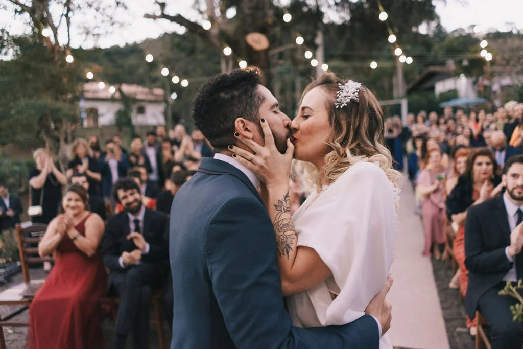 Um casal se beijando durante a cerimônia de casamento ao ar livre, com convidados ao fundo em comemoração.