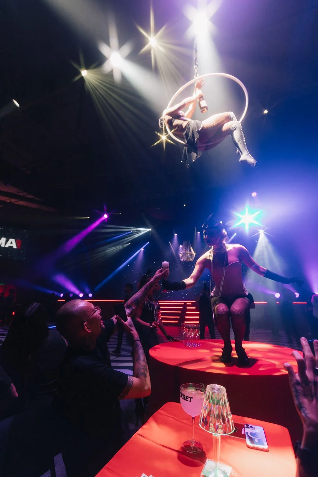 Show de acrobatas e bailarinas em um palco iluminado com luzes coloridas e telão de fundo, pessoas assistindo e fotografando a apresentação.