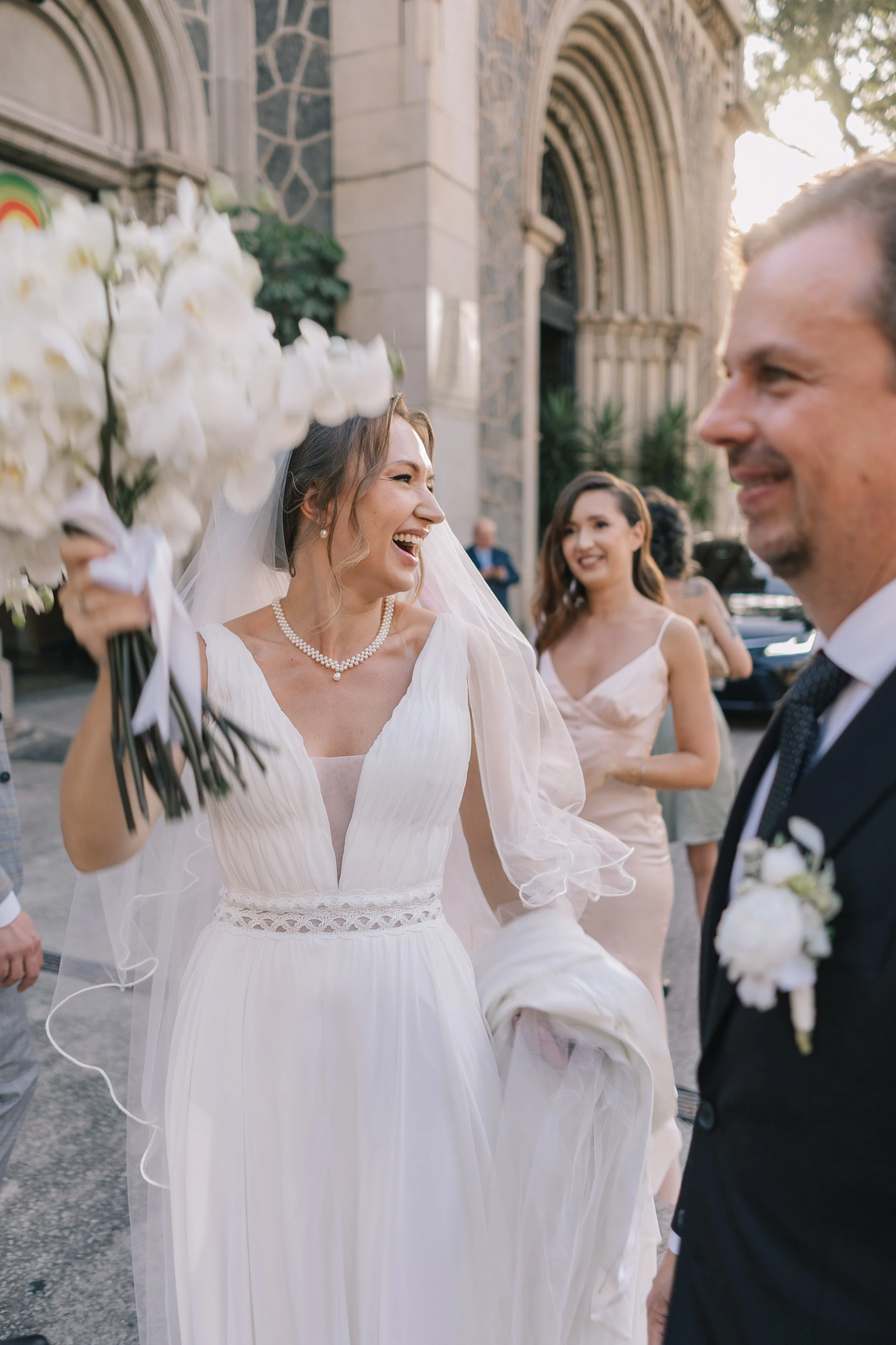 Noite de casamento ao ar livre com uma noiva sorridente segurando um buquê de flores brancas, ao lado de um noivo sorridente, com amigos ao fundo na entrada de uma igreja de pedra.