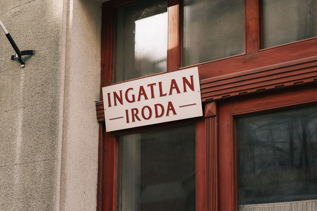 Placa de sinalização em janela de madeira com texto em húngaro e português, indicando um imóvel disponível para locação.