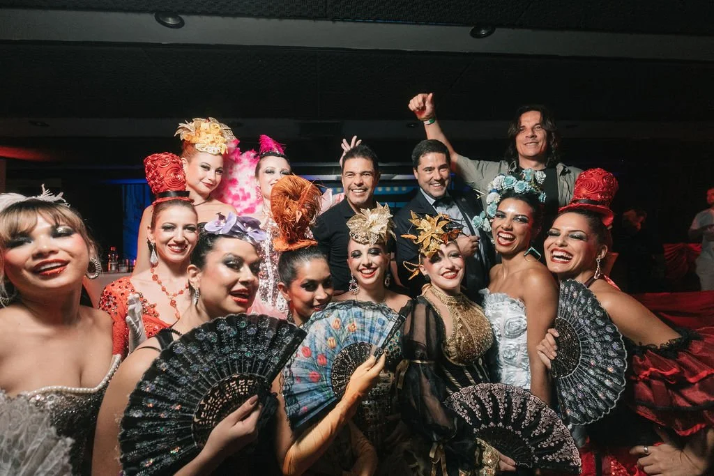 Grupo de pessoas vestidas com roupas de teatro ou fantasia, sorrindo e segurando leques, em um evento animado.