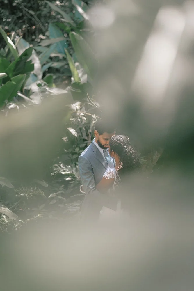 Casal de noivos abraçando-se em meio à vegetação densa, com folhas ao redor, durante um ensaio fotográfico ao ar livre.