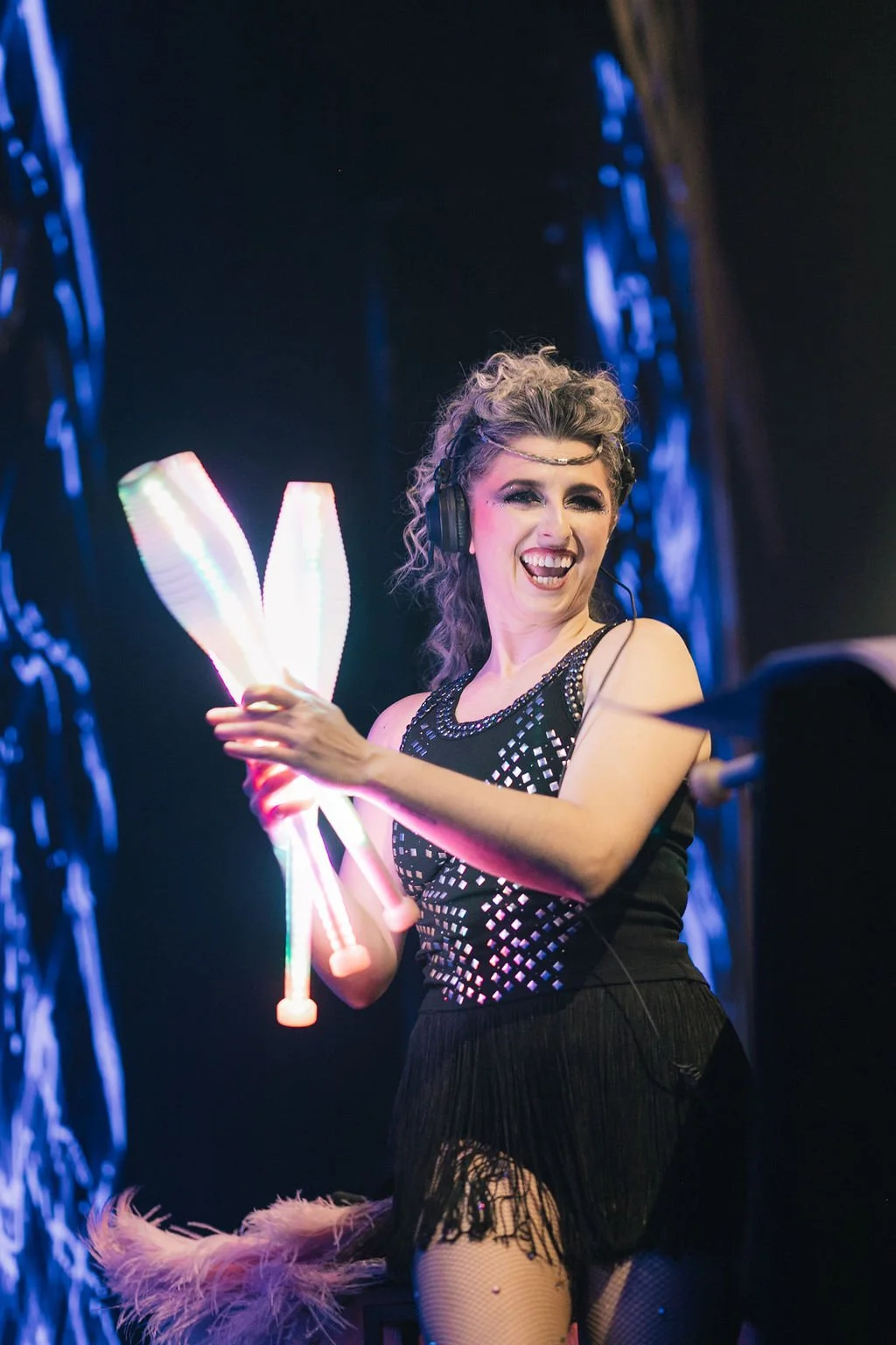 Mulher sorrindo, usando óculos de proteção auricular, segurando dois objetos luminosos em forma de V, com roupas de show brilhantes, em um palco com iluminação azul.