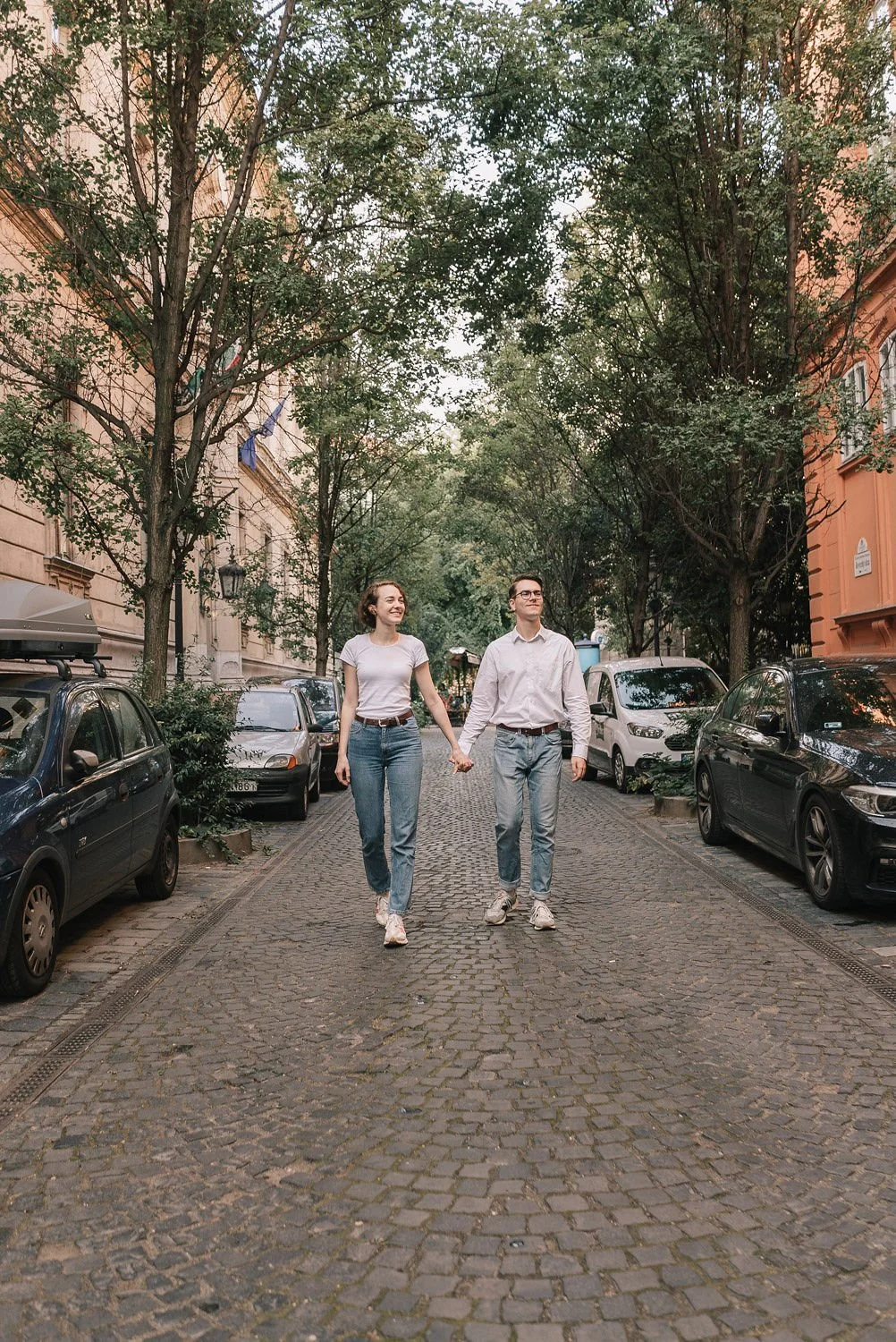 Casal jovem andando de mãos dadas na rua de paralelepípedo em uma rua arborizada, ao lado de carros estacionados.