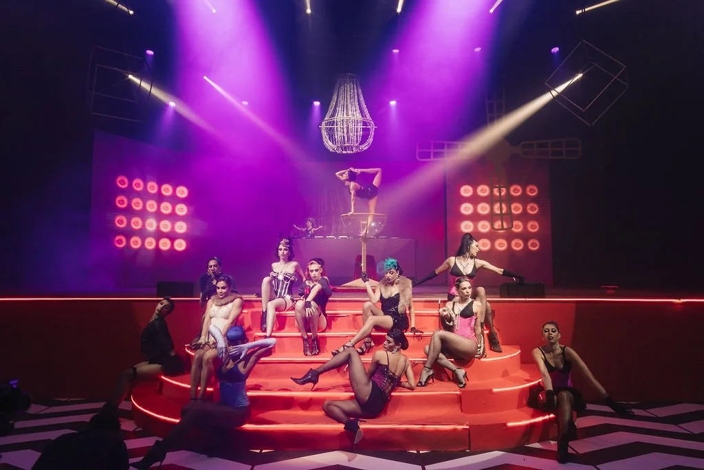 Cenário de teatro com várias pessoas em roupas de show, iluminação colorida, um chandelier no teto e uma escada circular com várias etapas, com pessoas posando e um DJ ao fundo.