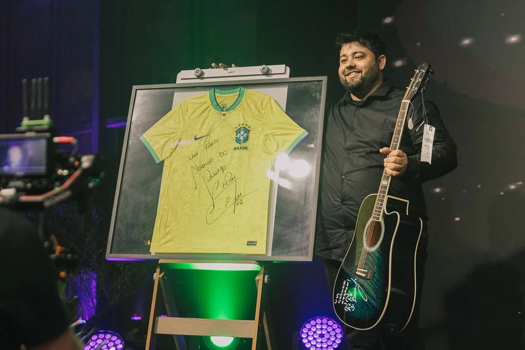 Homem segurando violão ao lado de quadro com uma camiseta amarela autografada da seleção brasileira de futebol.
