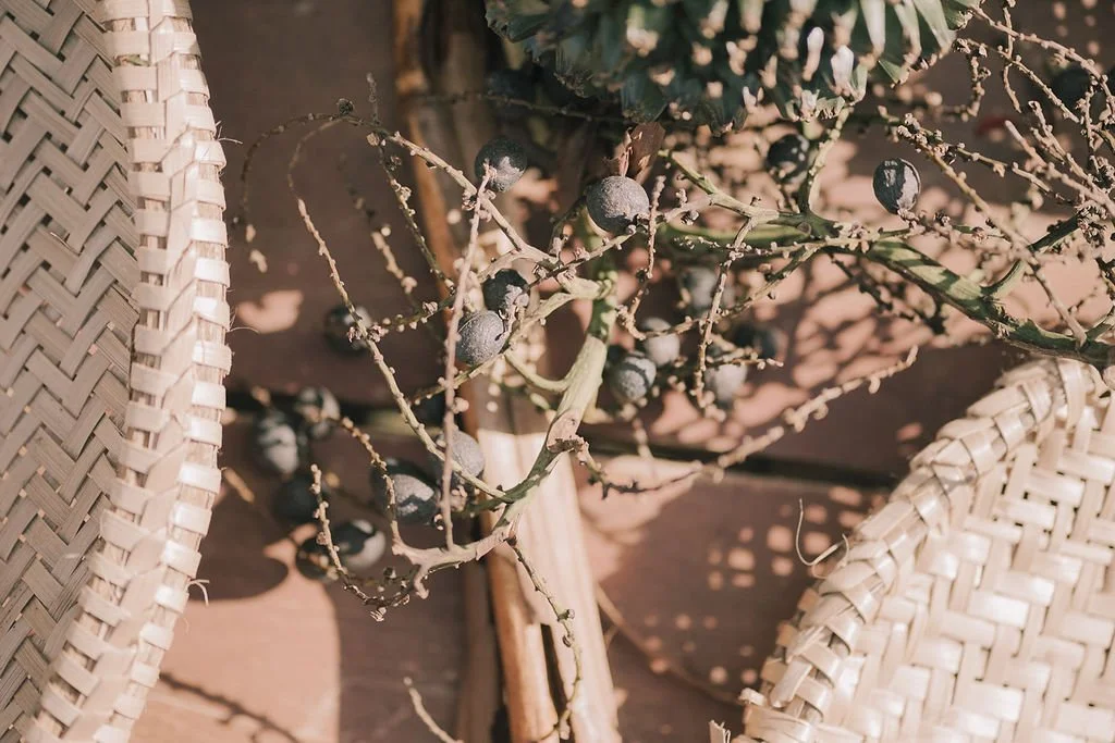 Galho seco com algumas frutas escuras e folhas, ao lado de cadeiras de vime, em uma mesa de madeira.
