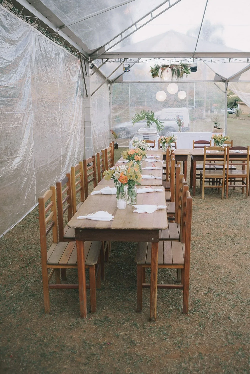 Mesa de madeira decorada com flores e talheres, sob uma estrutura de tenda para evento ao ar livre.