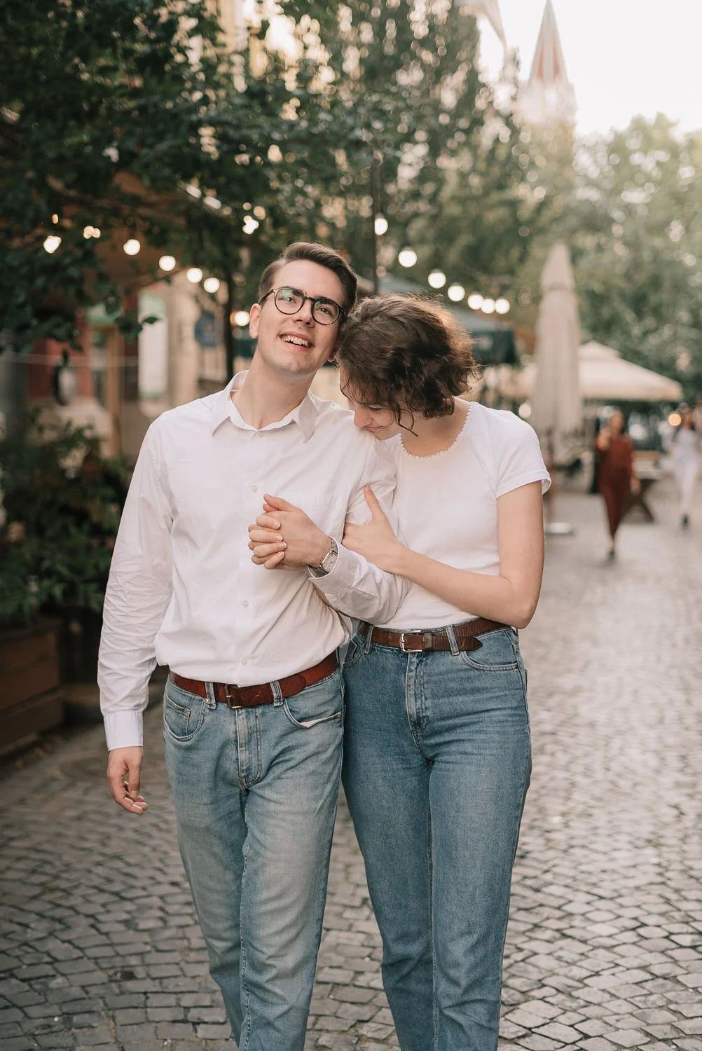 Um casal caminhando de mãos dadas na rua, sorrindo e se divertindo, com iluminação de luzes decorativas ao fundo em um ambiente ao ar livre.