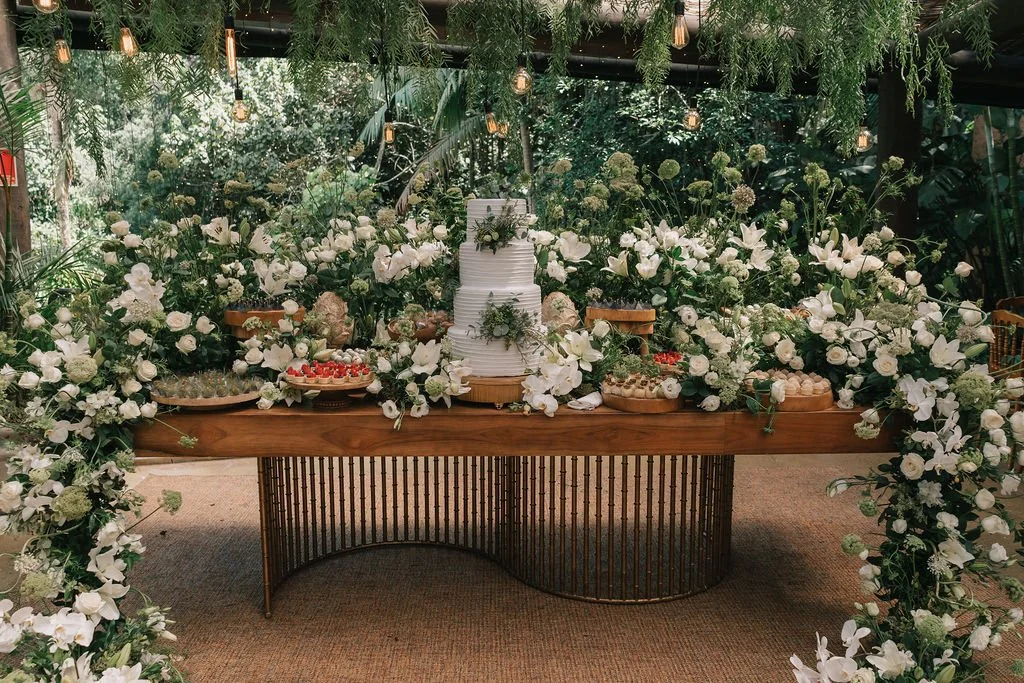 Mesa decorada com flores brancas e um bolo de casamento no centro, em um ambiente com iluminação suave e vegetação ao fundo.