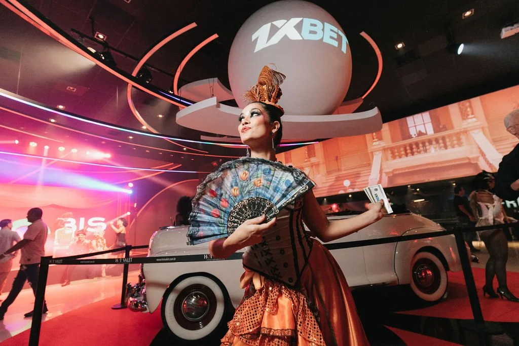 Mulher vestida em traje de dominating, com uma cabeça de touro decorativa, segurando um leque e dinheiro, em evento com iluminação colorida e um carro clássico ao fundo, em um ambiente de festa.