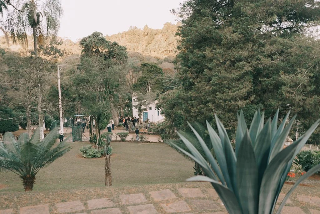 Vista de um jardim com árvores e plantas, com algumas pessoas ao fundo, próximo a uma estrutura de igreja ou capela em uma área rodeada por natureza com montanhas ao fundo.