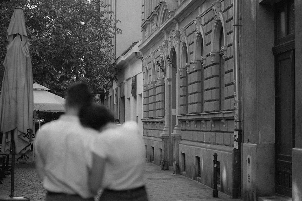 Duas pessoas de costas caminhando em uma rua de cidade antiga, ao lado de um prédio com detalhes arquitetônicos clássicos.