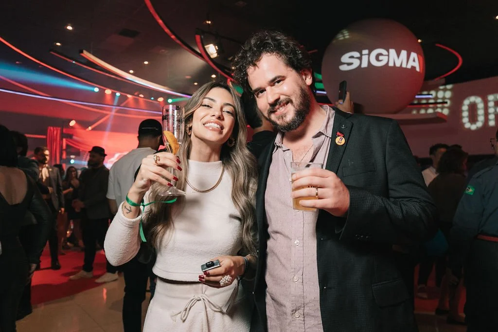Homens e mulheres festejando em uma festa com iluminação colorida, uma pessoa com vestido branco e outra com blazer preto segurando copos de bebida.