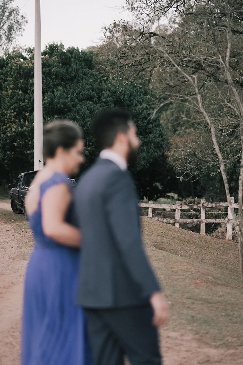 Casal vestindo roupas formais caminhando ao ar livre, com árvores e cerca de madeira ao fundo, e um carro preto estacionado ao lado.