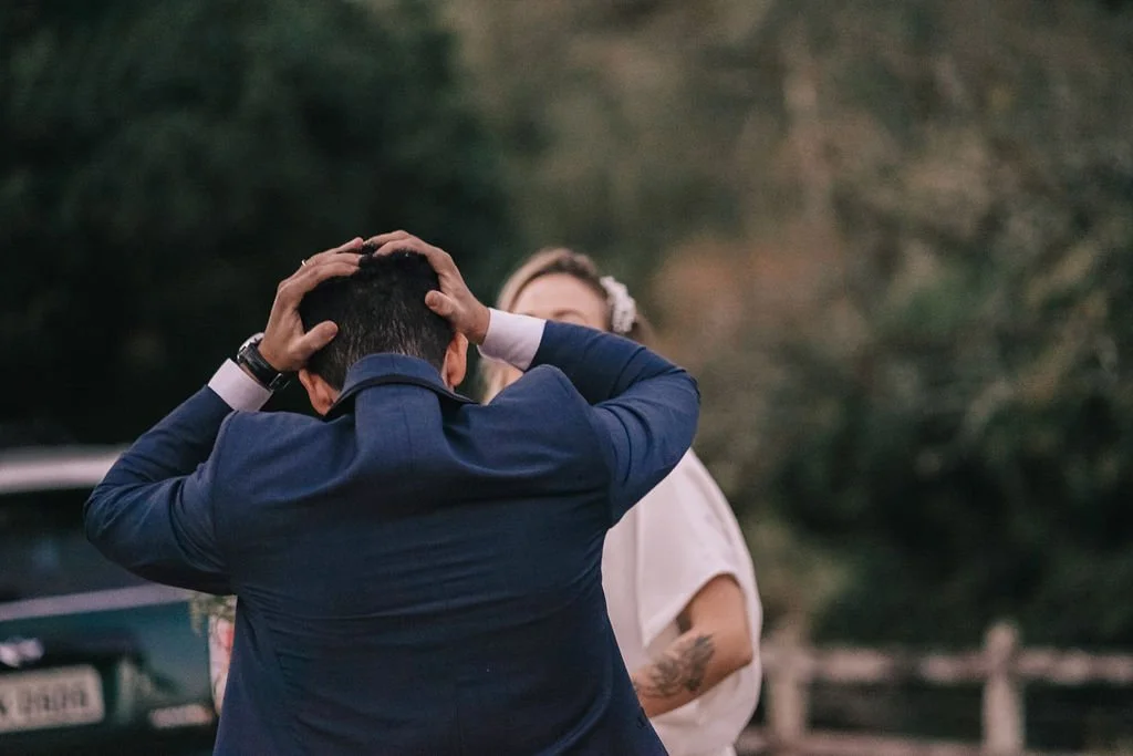 Homem em traje formal assustado ou surpreso, segurando a cabeça com as mãos, enquanto uma mulher com cabelo encaracolado e vestida de branco olha para ele ao fundo, ao ar livre.
