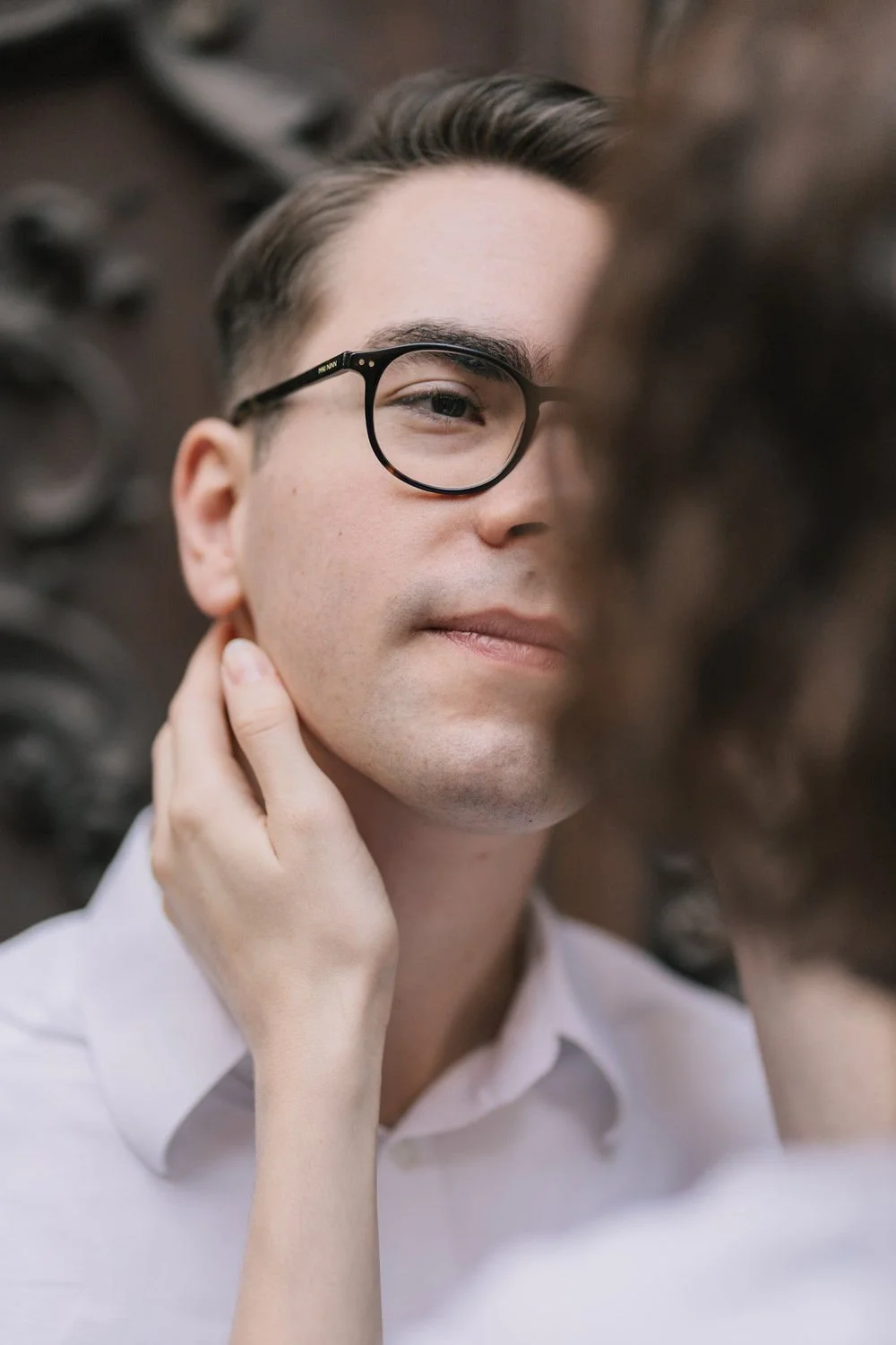 Homem com óculos escuros, cabelo penteado para trás, usando camisa branca, ao lado de uma mulher com cabelo castanho escuro, em uma expressão de carinho.