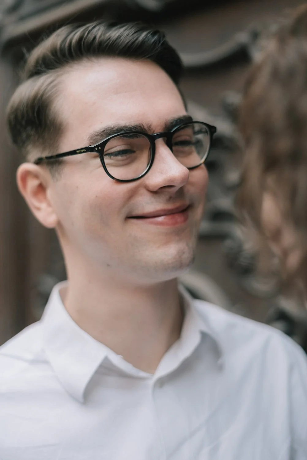 Homem sorridente usando óculos de armação preta e camisa branca, com fundo desfocado.