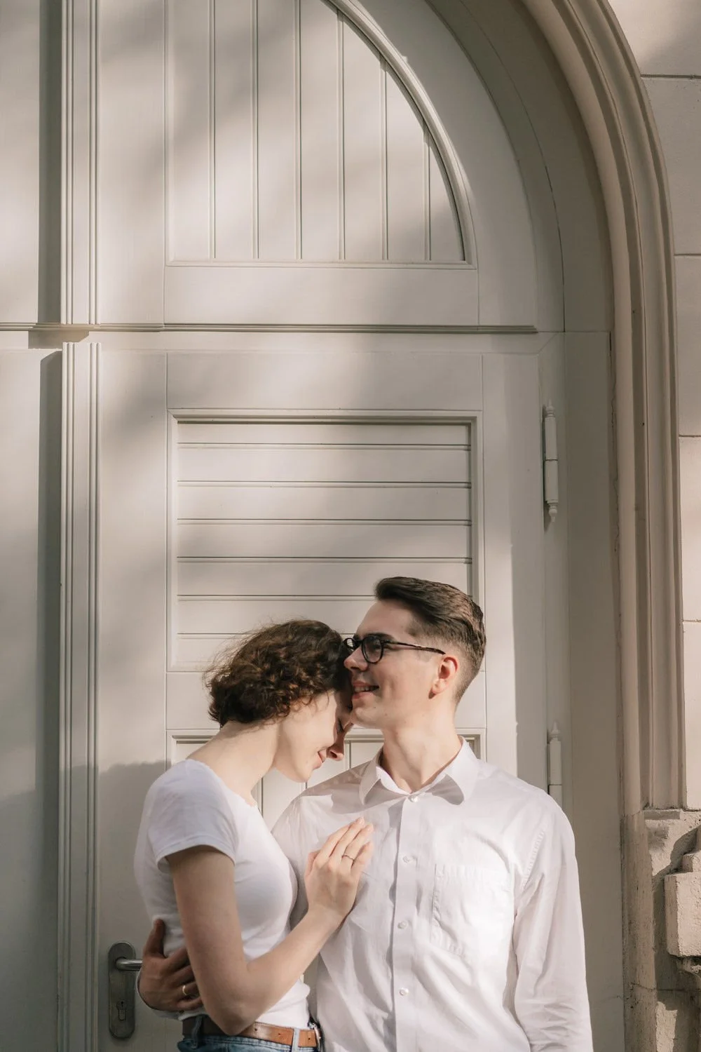 Um casal jovem com roupas brancas, brincando e sorrindo de cabeça encostada na porta branca de uma casa ou edifício, ao pôr do sol.