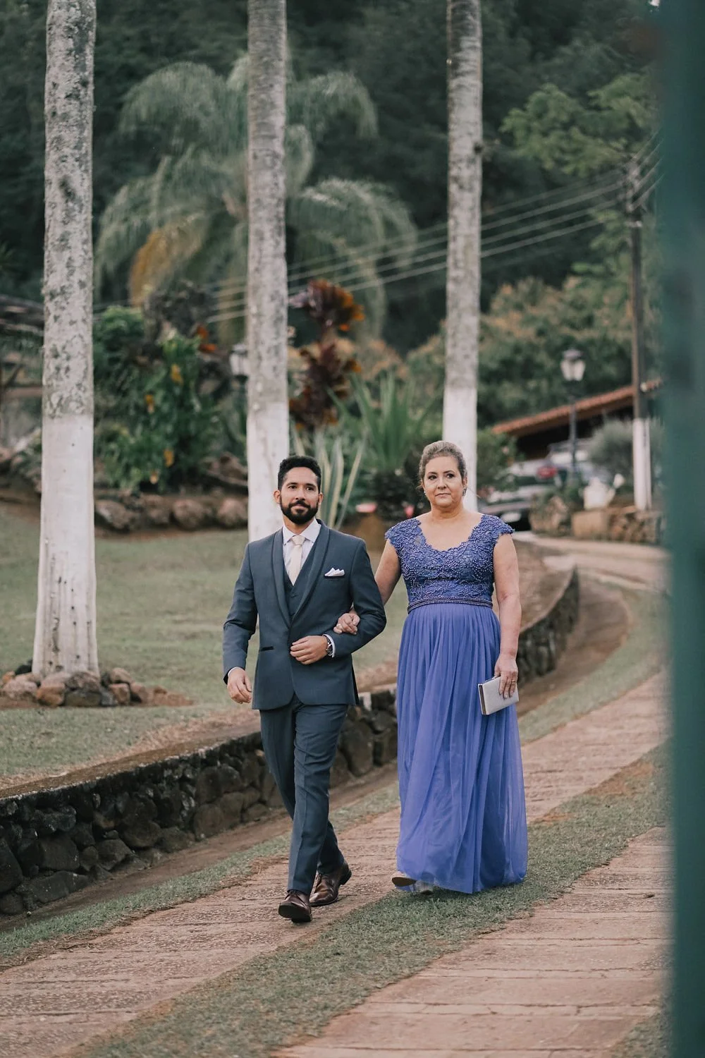 Homem e mulher vestidos formalmente caminhando ao ar livre, com árvores e vegetação ao fundo