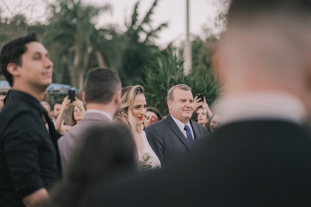 Casal sorridente em evento ao ar livre, com pessoas ao redor, algumas tirando fotos.