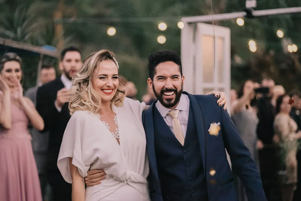Casal sorridente em cerimônia de casamento, com convidados ao fundo. Ela usa vestido claro e ele terno azul escuro.