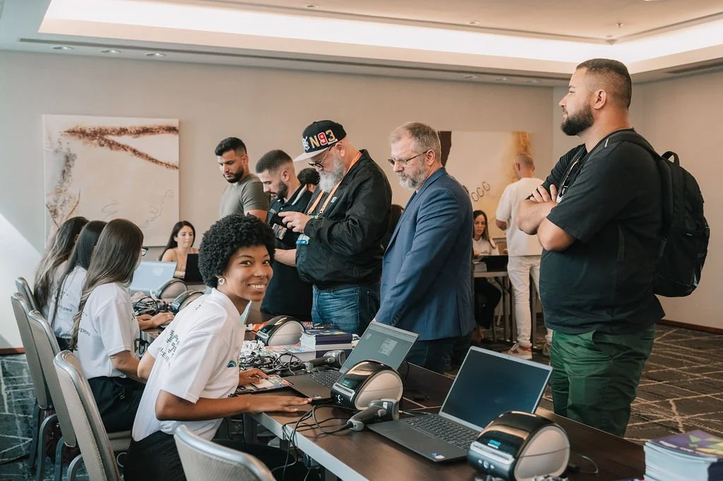 Pessoas em uma sala de treinamento ou conferência, com alguns usando computadores, enquanto outros conversam ou observam, incluindo uma mulher sorridente sentada na frente.