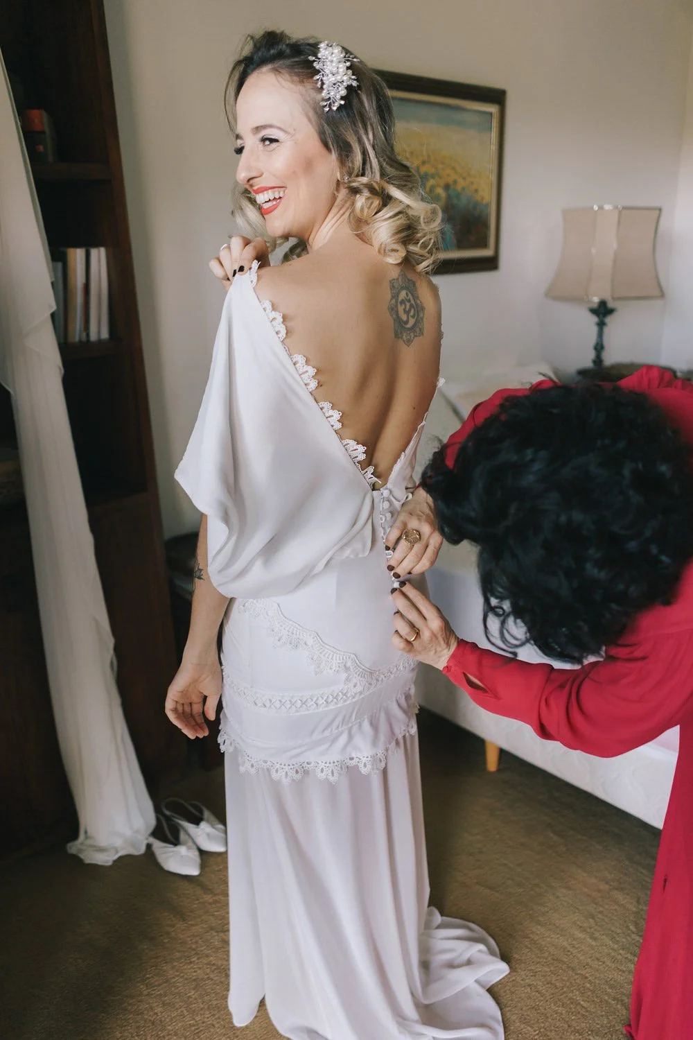 Mulher feliz de vestido de noiva branco sendo ajustada por outra pessoa no quarto.