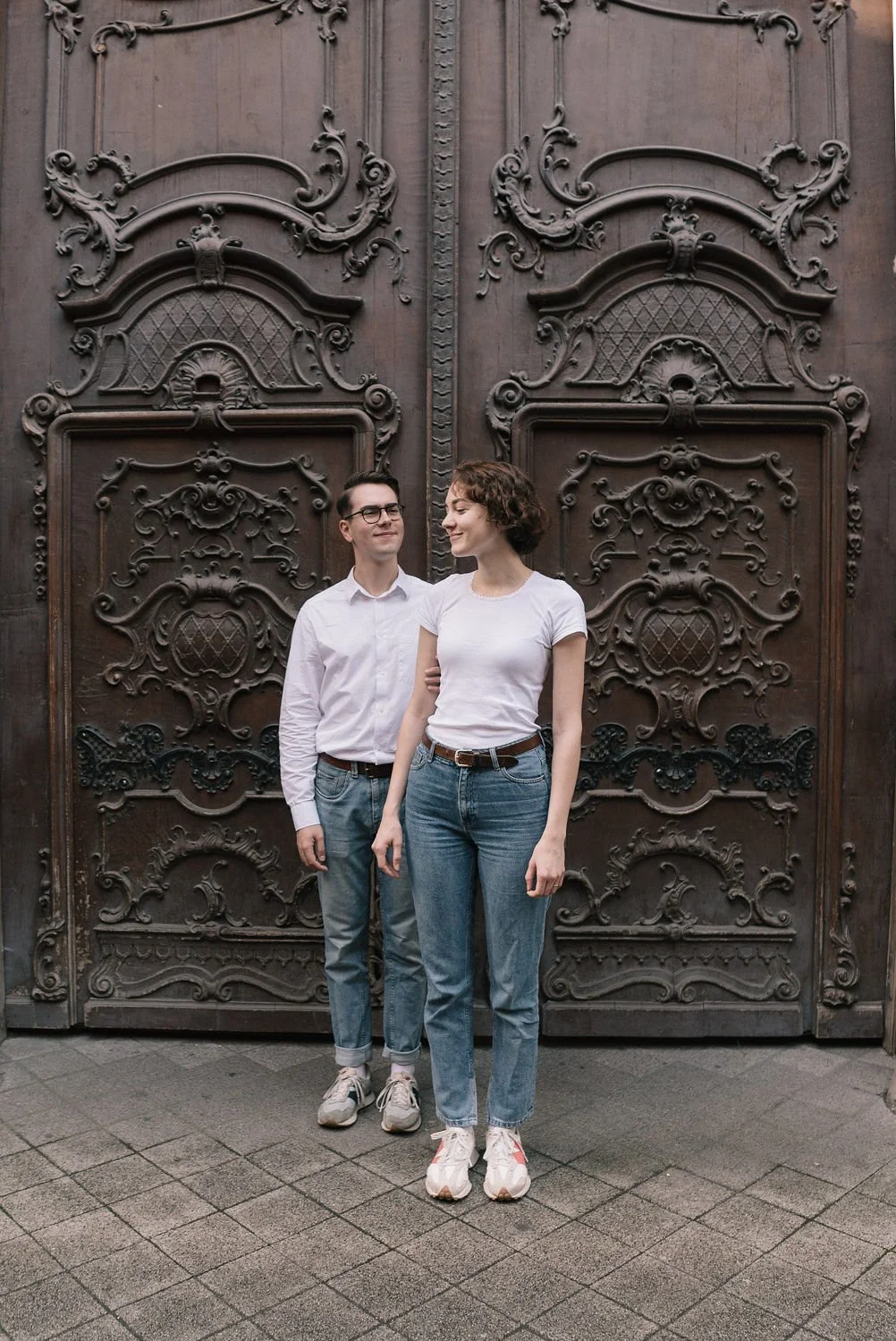 Homens e mulher de camiseta branca e jeans posando na frente de uma porta de madeira ornamental.