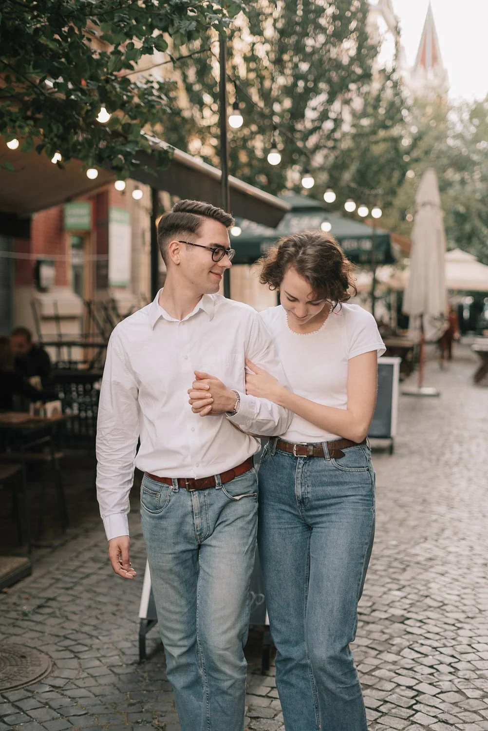 Um casal caminhando de mãos dadas na rua ao pôr do sol, com árvores e luzes penduradas acima, em um ambiente ao ar livre.