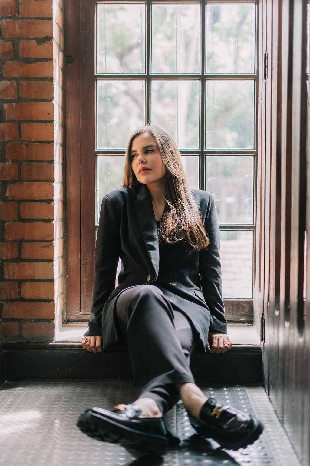 Mulher jovem com cabelo castanho claro, vestindo terno preto, sentada no chão próximo a uma janela com moldura de madeira, dentro de um ambiente com parede de tijolos e piso de borracha, olhando pensativa para o lado.
