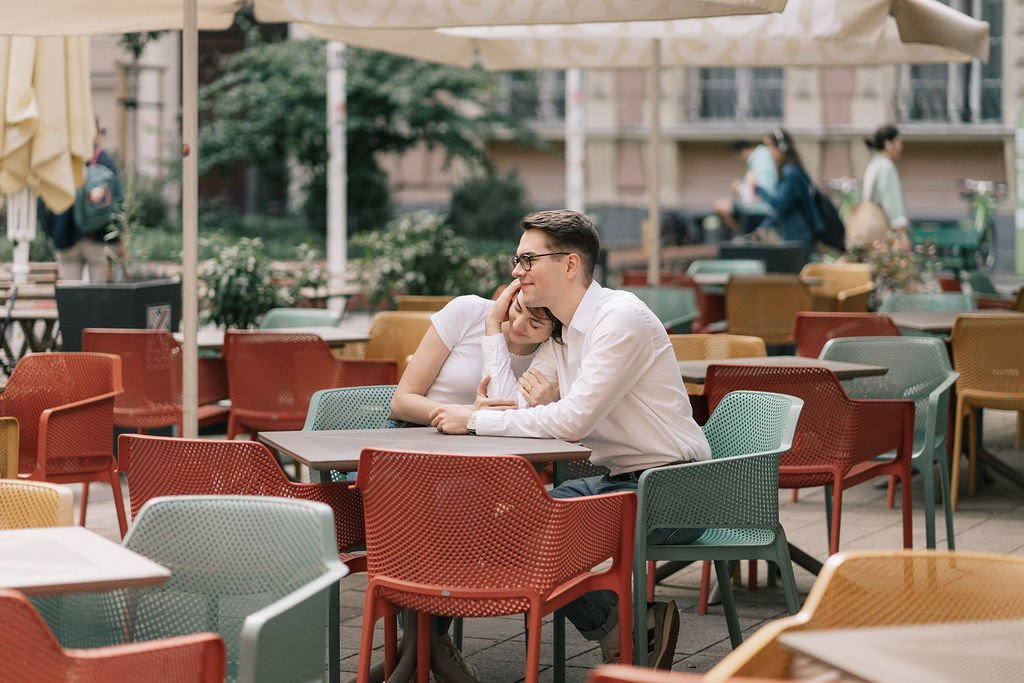 Casal sentado em uma mesa ao ar livre de um café, compartilhando um momento de carinho, com cadeiras coloridas ao redor e uma cobertura acima, enquanto outras pessoas caminham ao fundo.