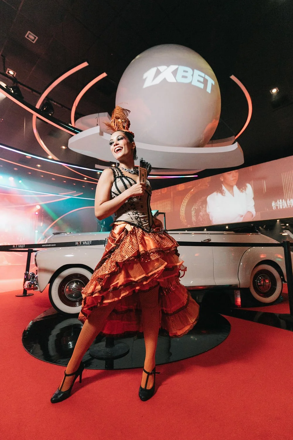 Mulher sorridente vestida com roupa de estilo vintage e acessório de cabeça brilhante, segurando notas de dinheiro, em evento com decoração luxuosa, carro antigo e projeção de logo de site de apostas.