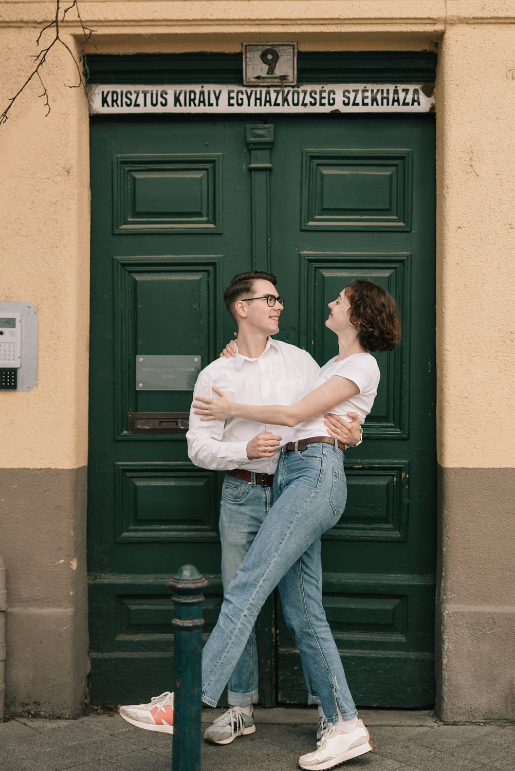 Casal jovem se abraçando em frente a uma porta verde com um símbolo na parte superior e uma placa ao lado.