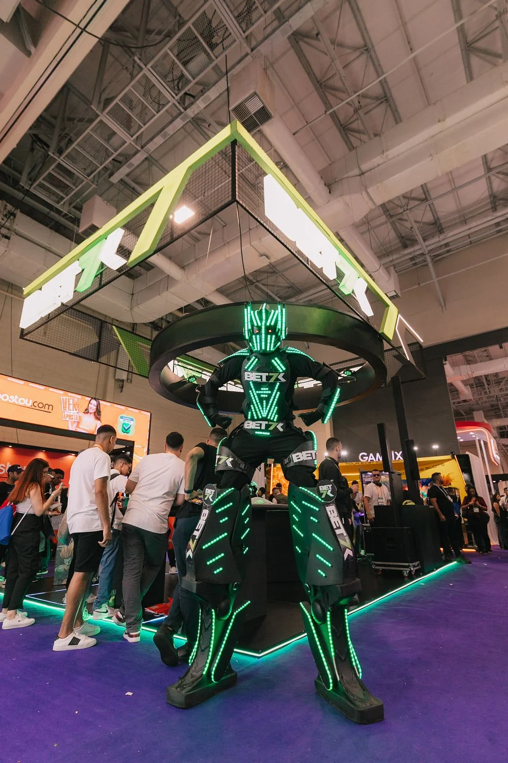 Robô de aparência futurista com detalhes em LED verde na cabine de um expositor na feira, com pessoas ao redor observando.