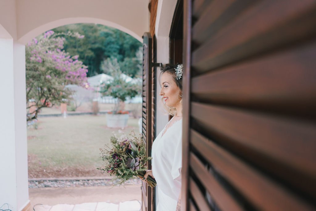 Mulher sorridente segurando um buquê de flores, vista de perfil através de uma janela de madeira, ao lado de uma porta de entrada, com jardim ao fundo.