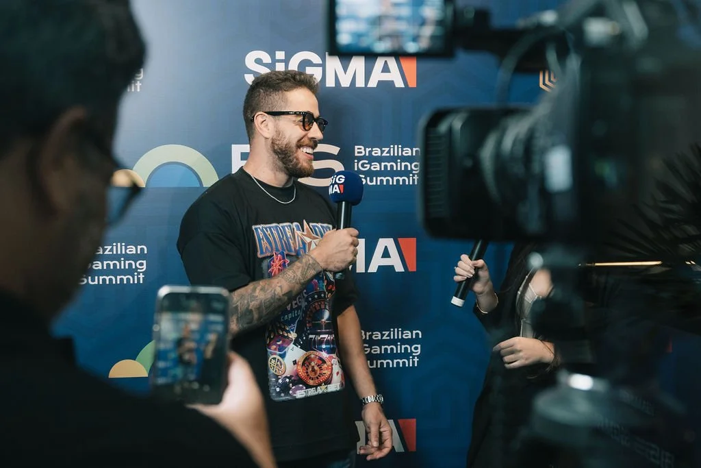 Homem com barba e tatuagens sendo entrevistado em evento de jogos no Brasil, com fundo azul e logo do Brazilian iGaming Summit.