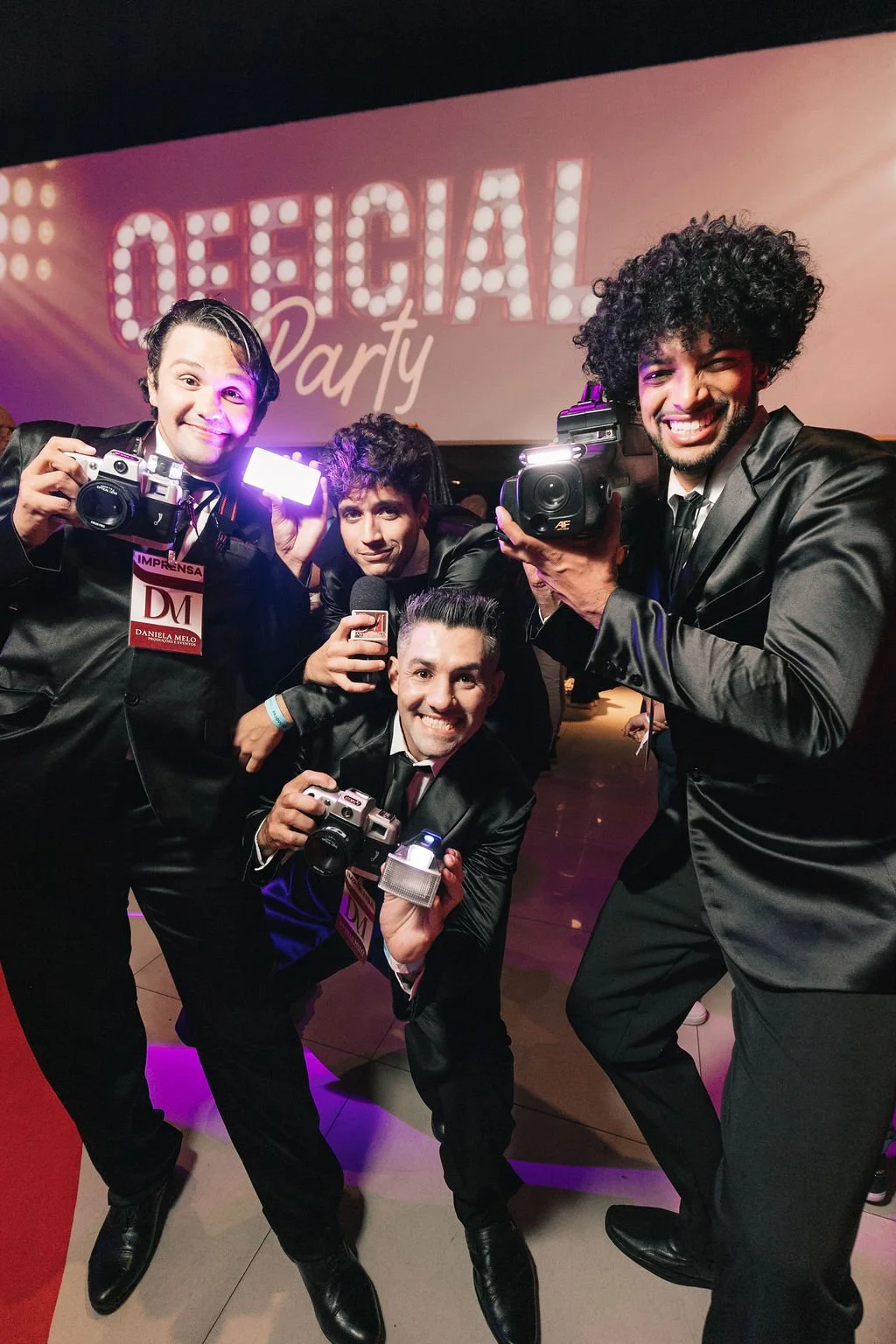 Grupo de quatro homens sorridentes em traje formal, segurando câmeras e microfone em uma festa com luzes profissionais e uma tela ao fundo que diz 'Official Party'.