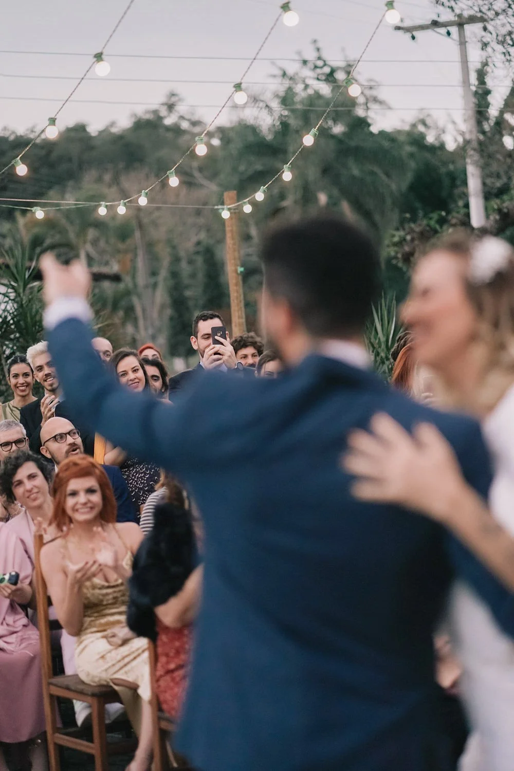 Casal dançando em celebração ao ar livre com alegria, cercado por convidados sorridentes, sob luzes decorativas penduradas no céu.