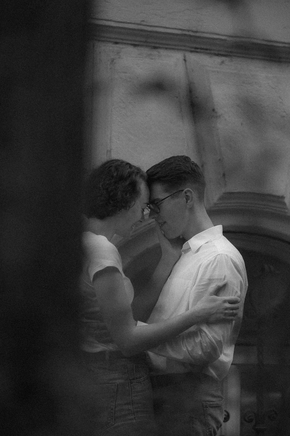 Imagem em preto e branco de um casal na ponta de seus corpos, com suas testas juntas, olhos fechados, tocando as faces um do outro. Eles estão em um ambiente interno, possivelmente uma igreja ou um local de cerimônia, com uma estrutura de arco ao fun