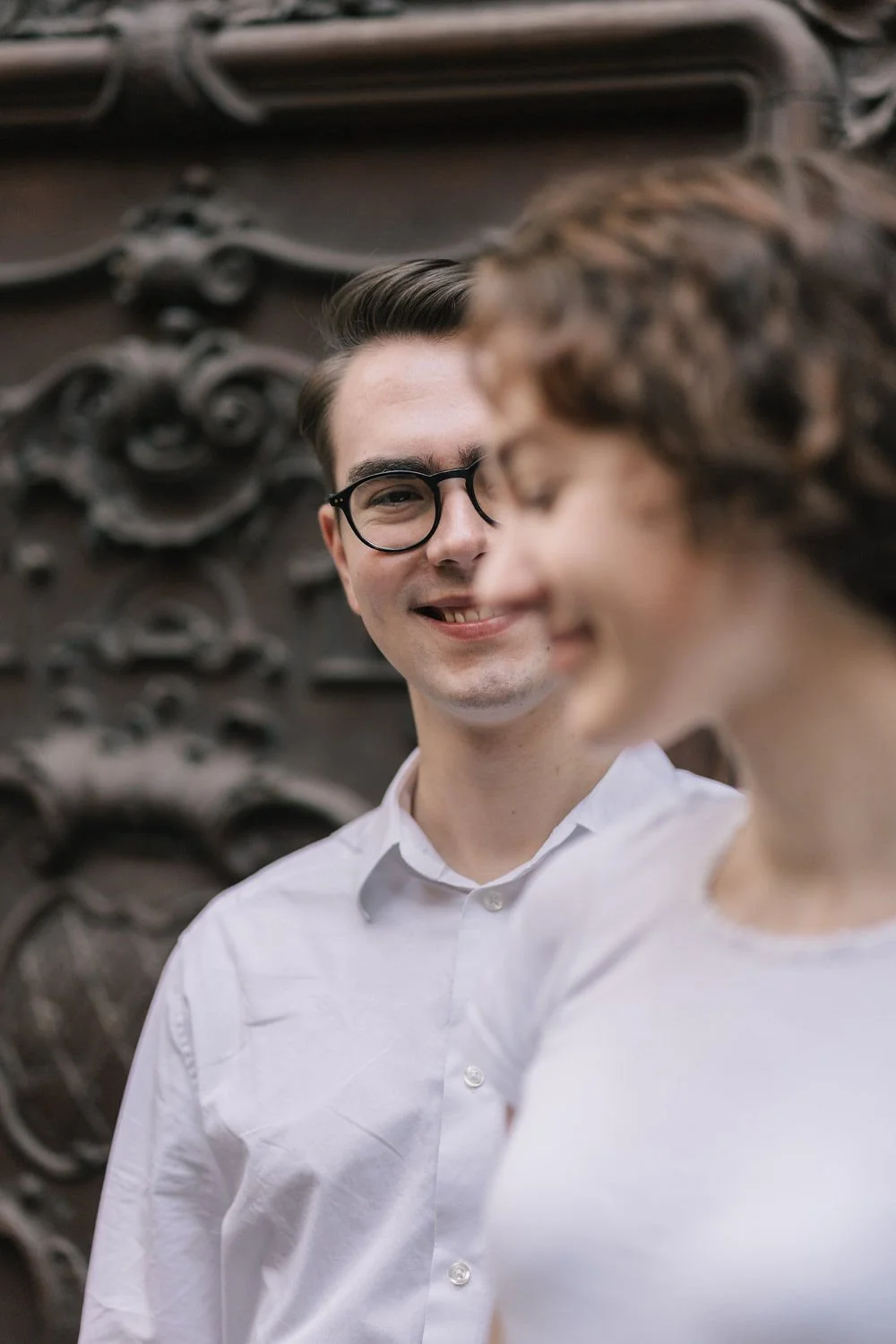 Homem sorrindo usando óculos de grau, ao lado de uma mulher, com fundo de relevo artístico em metal.