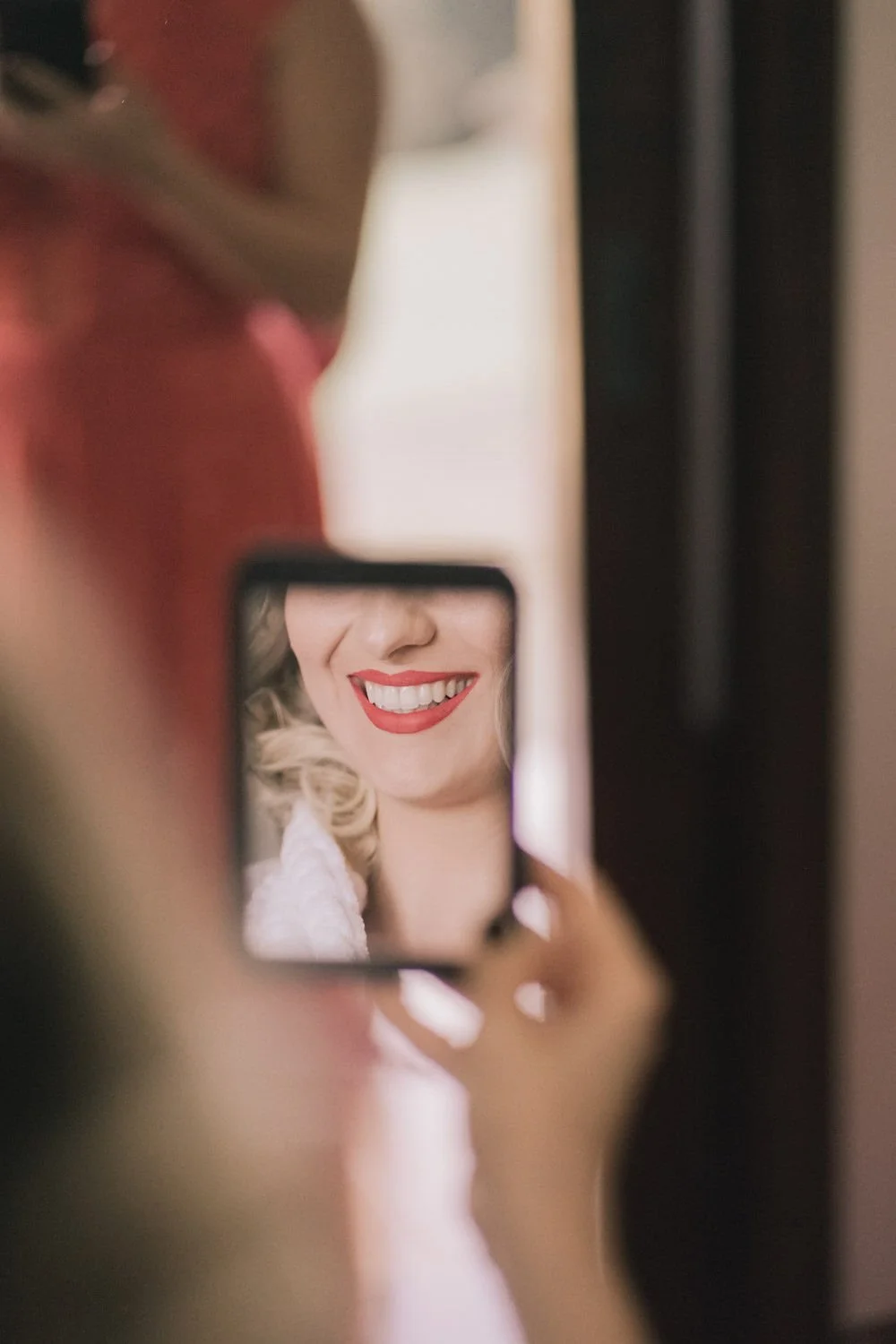 Pessoa segurando celular que captura seu sorriso com batom vermelho e cabelo cacheado, refletindo uma imagem sorridente.