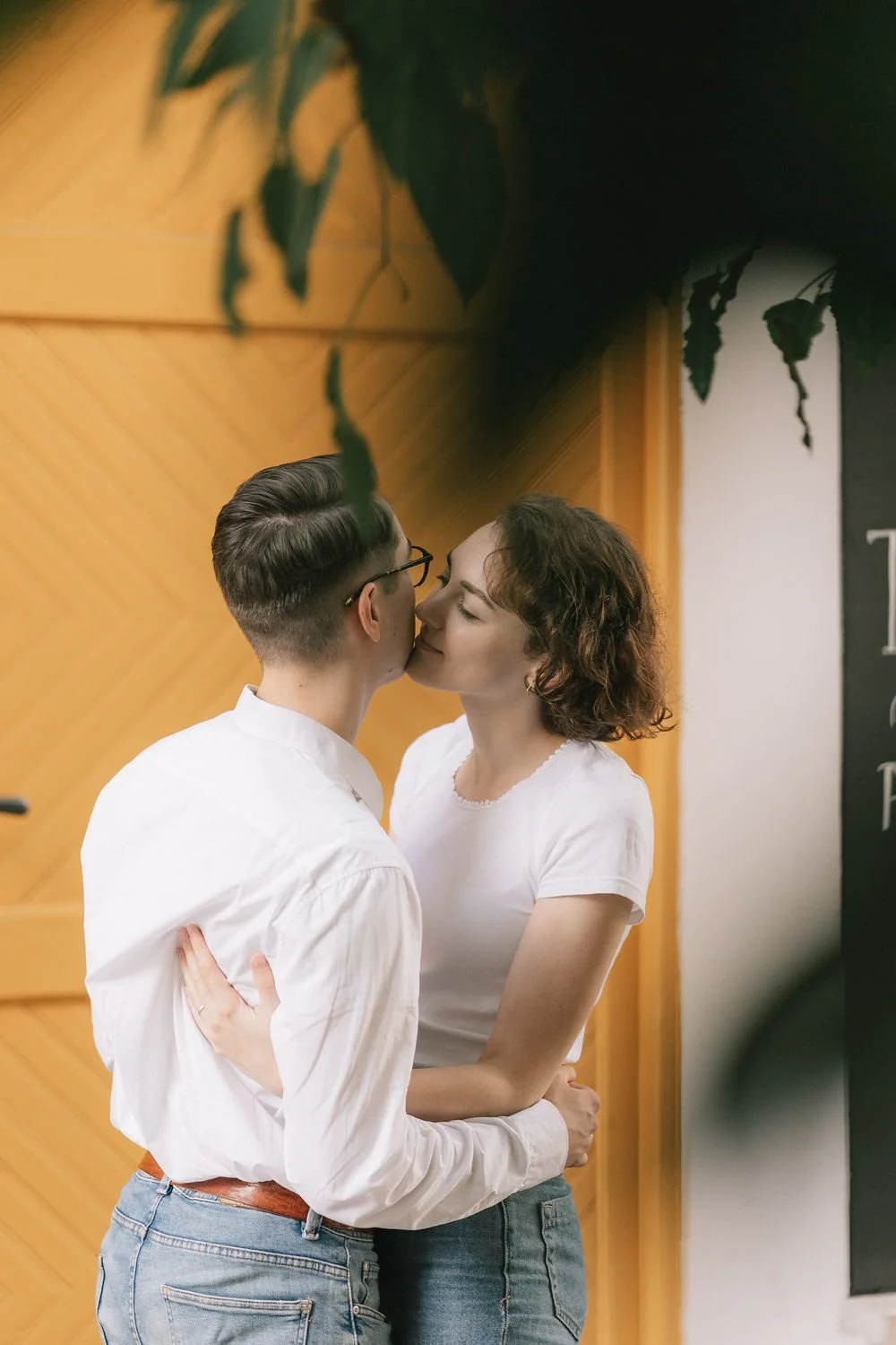 Casal jovem se beijando, de pé, em ambiente com parede de madeira e uma folha de planta visível na parte superior da imagem.