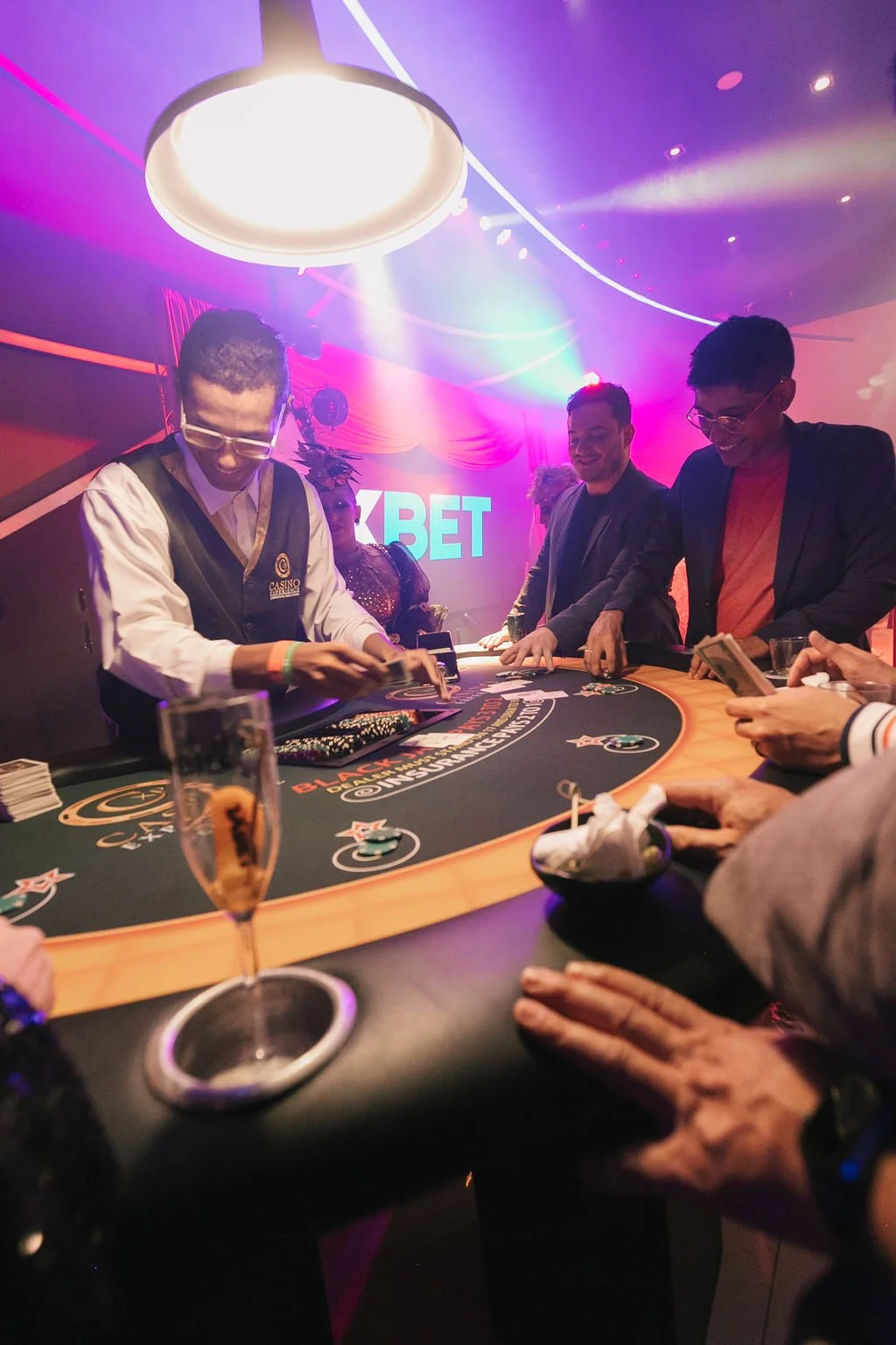 Pessoas jogando roleta em um cassino com iluminação colorida e uma tela ao fundo com escrita 'XBET'
