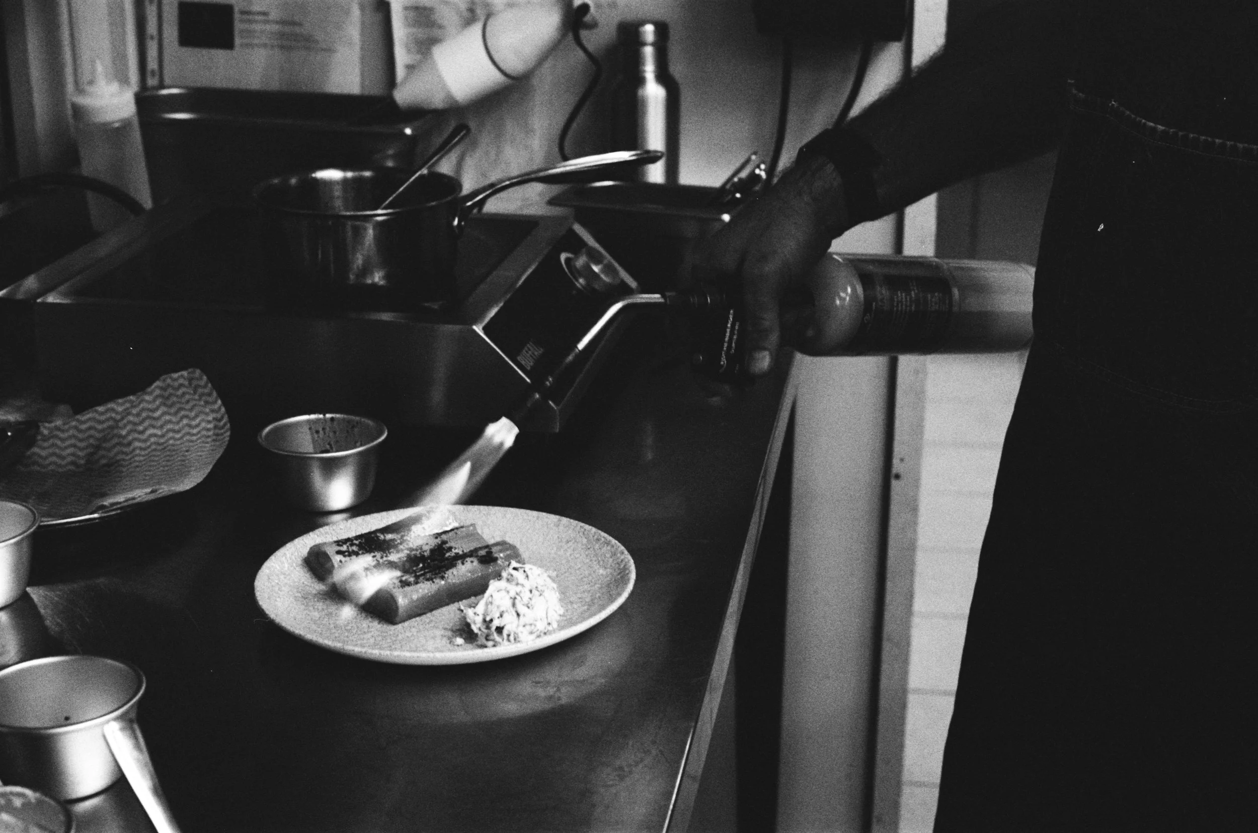 Person in black shirt and gloves preparing food on a stove, with a plate of rolled dishes and a dollop of condiment in the foreground.