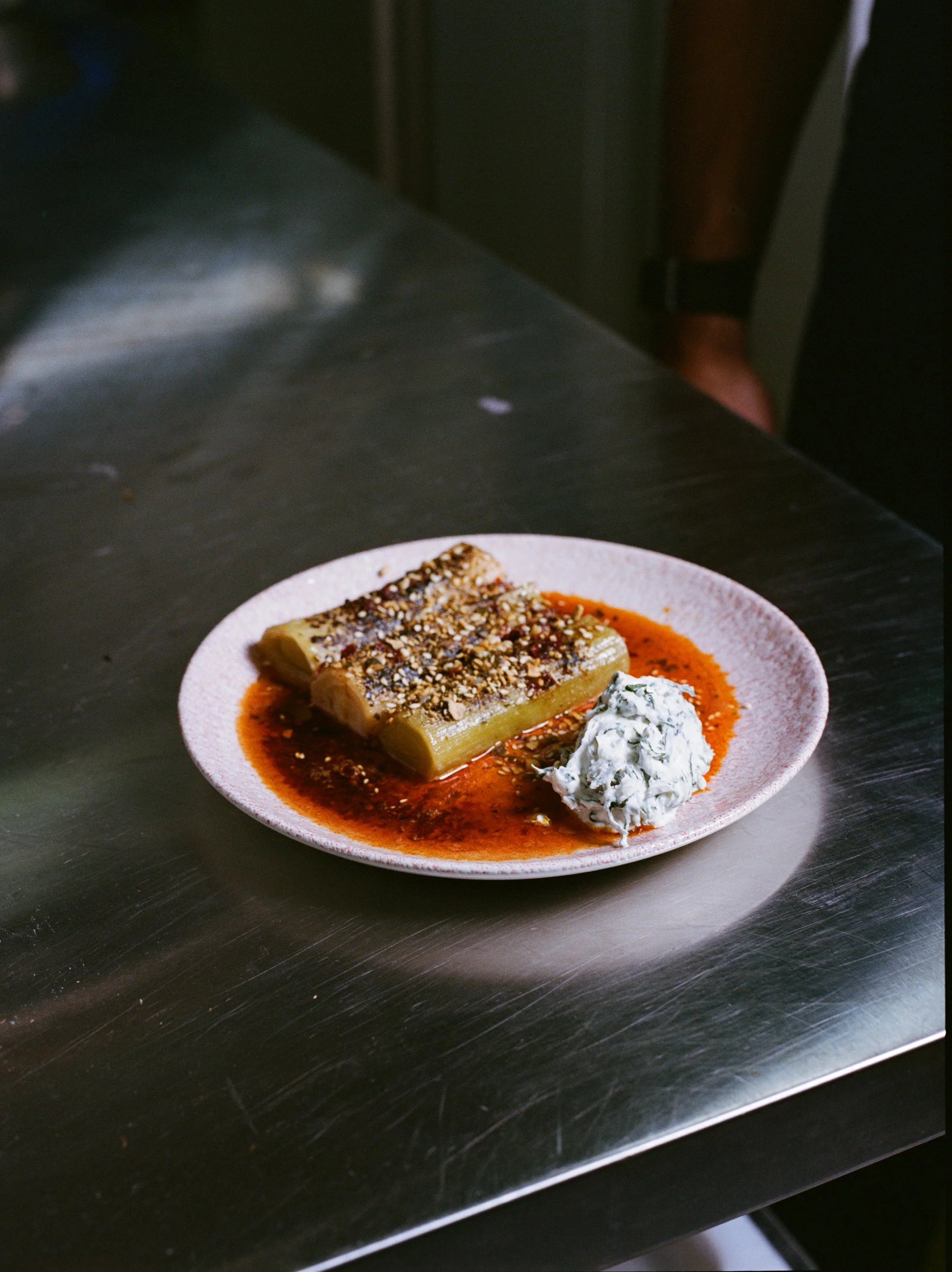 A white plate with stuffed green chili topped with sesame seeds and red sauce, with a dollop of creamy white sauce or dip next to it, on a stainless steel countertop.